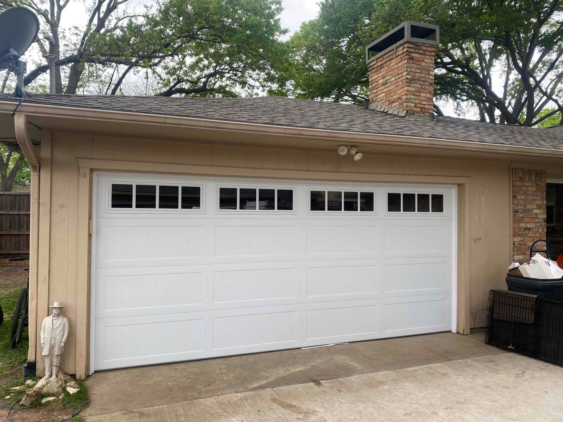 A white, four-panel residential garage door with top-row windows on a tan building with a brick chimney.
