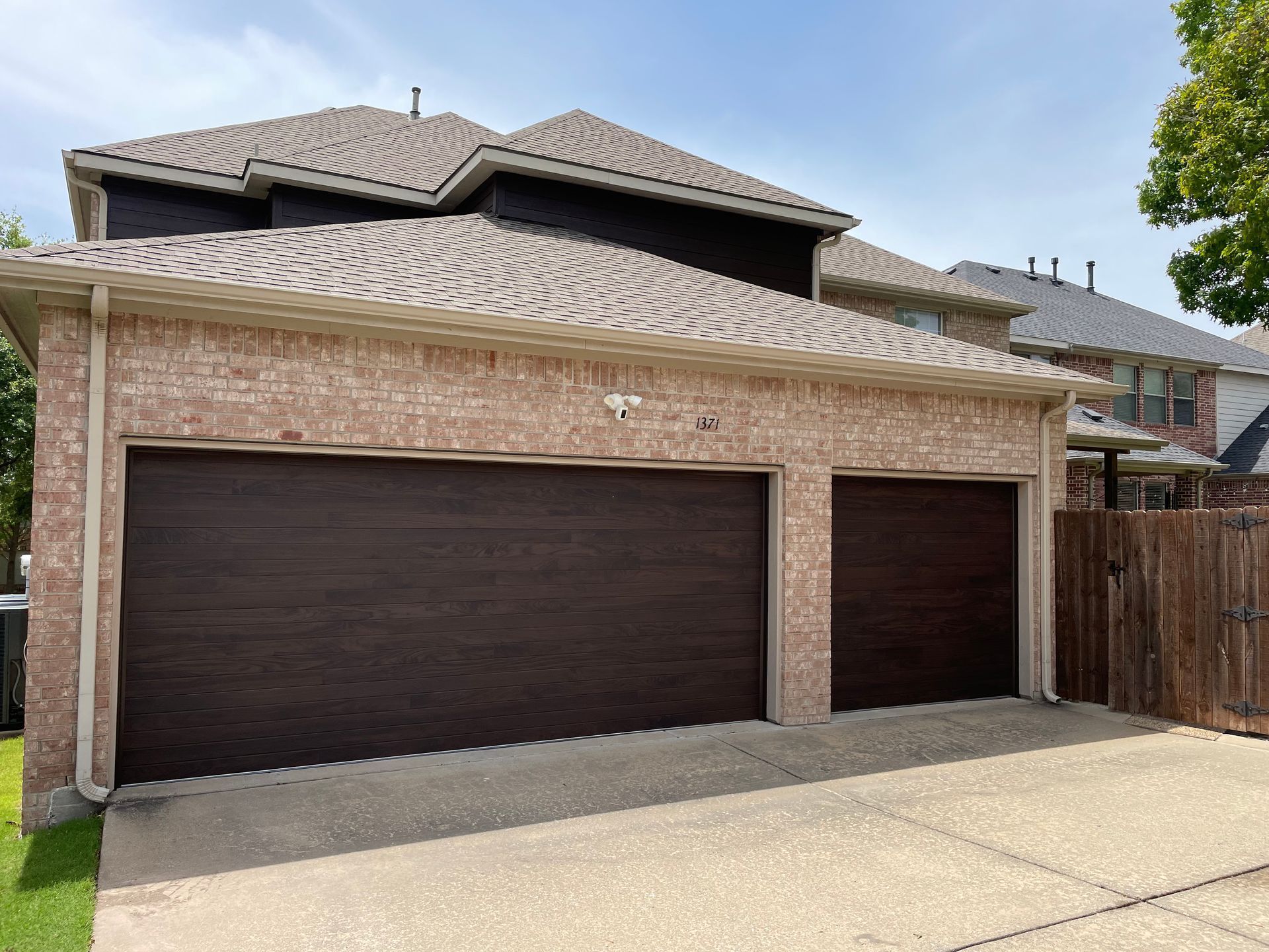 A house exterior featuring a beige brick facade, a multi-gabled roof, and two dark brown garage doors under a blue sky.
