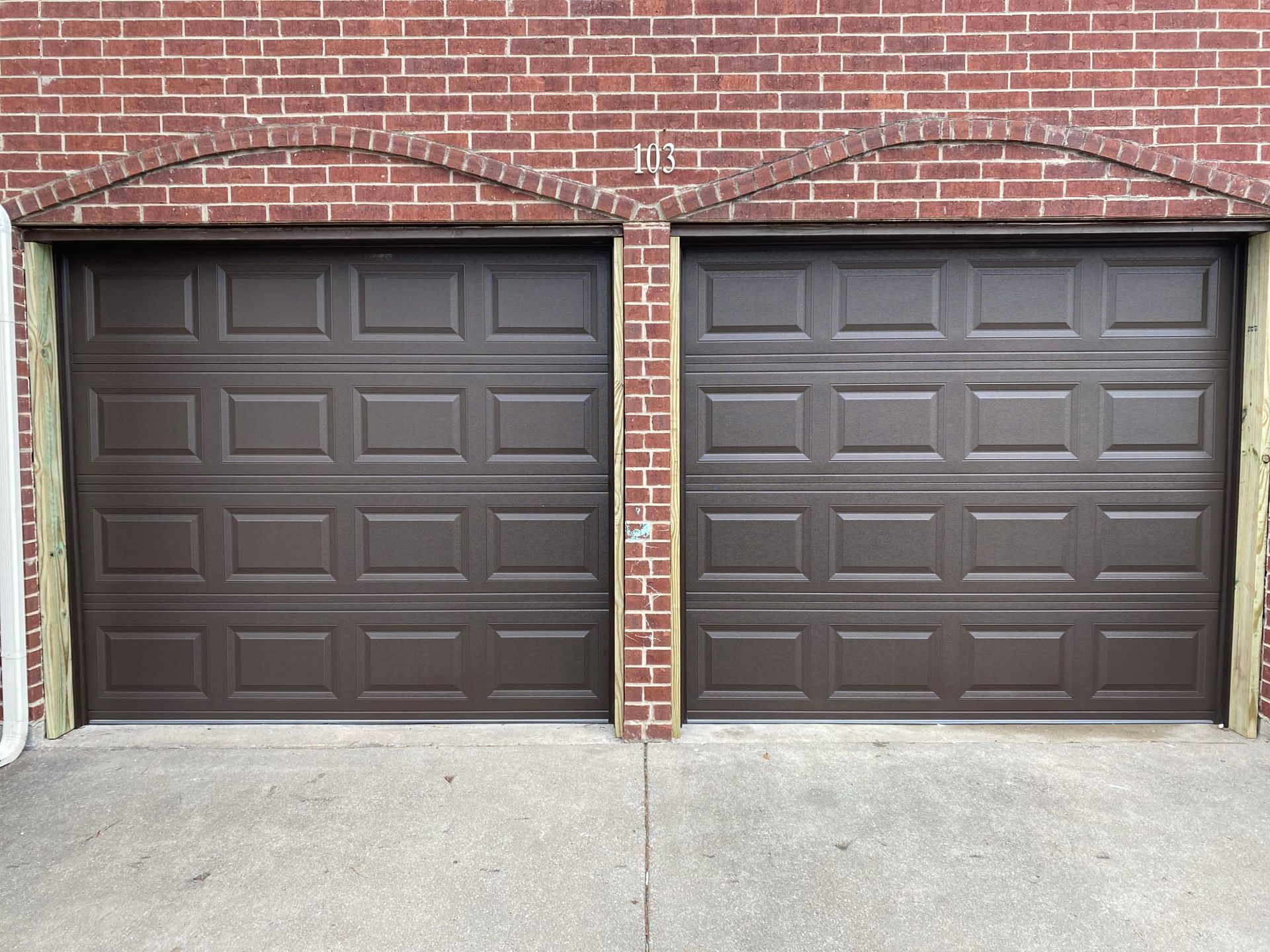 Two brown, paneled garage doors side-by-side set into a brick exterior wall.
