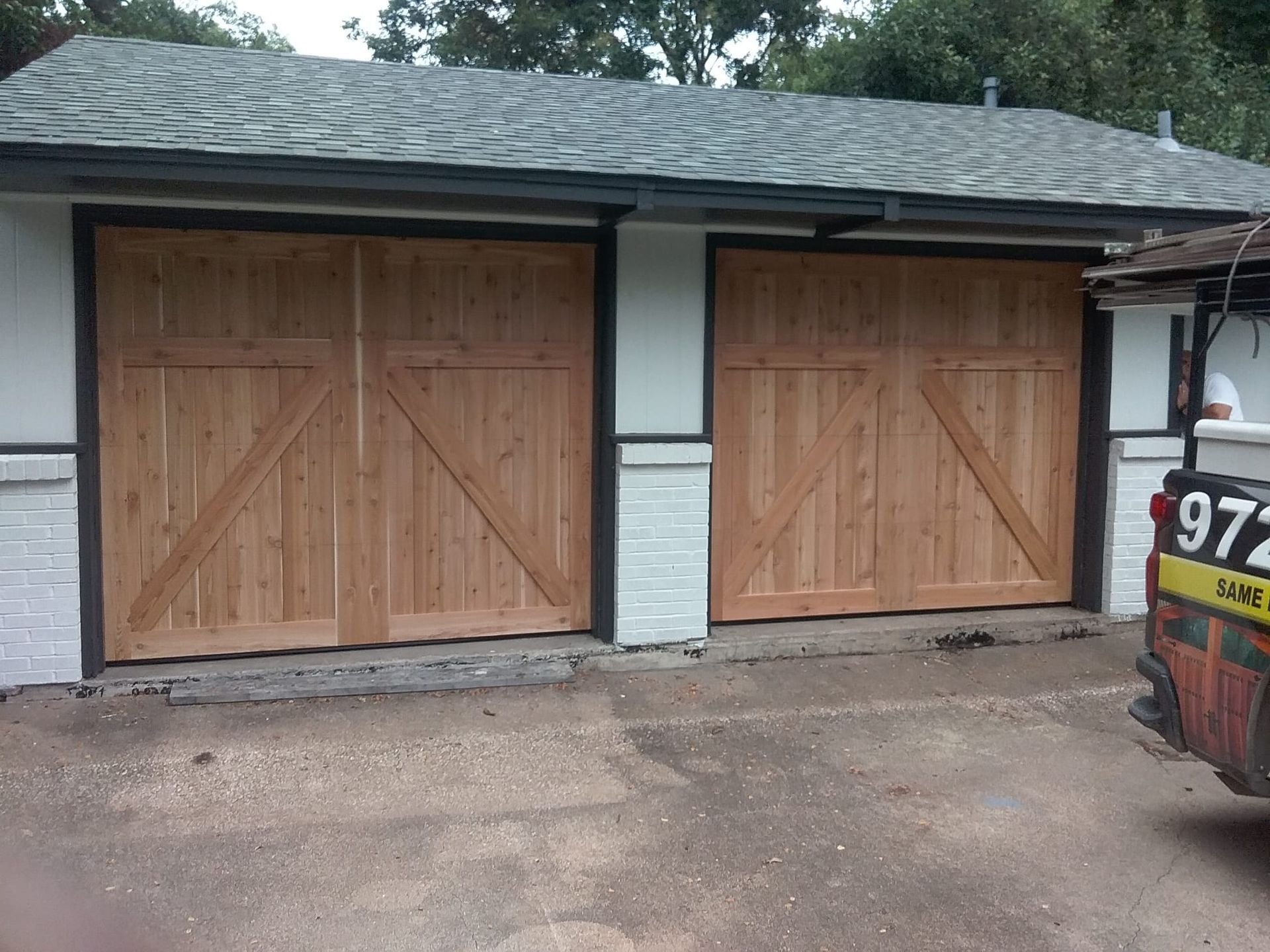 Two wooden carriage-style garage doors with diagonal bracing installed in a white building with a gray shingled roof.