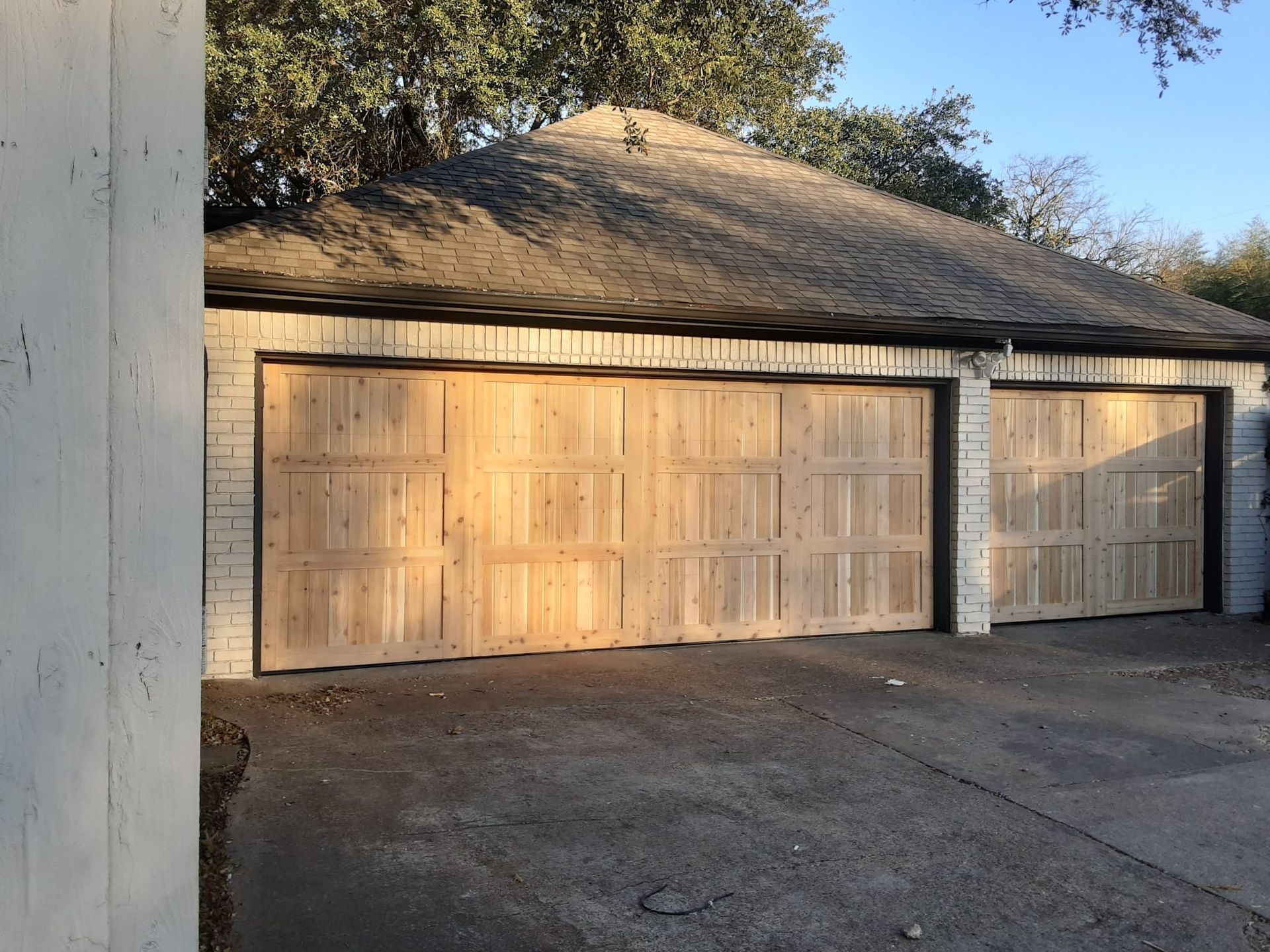A detached garage with two light-wood paneled doors, white brick siding, and a dark shingled hip roof.