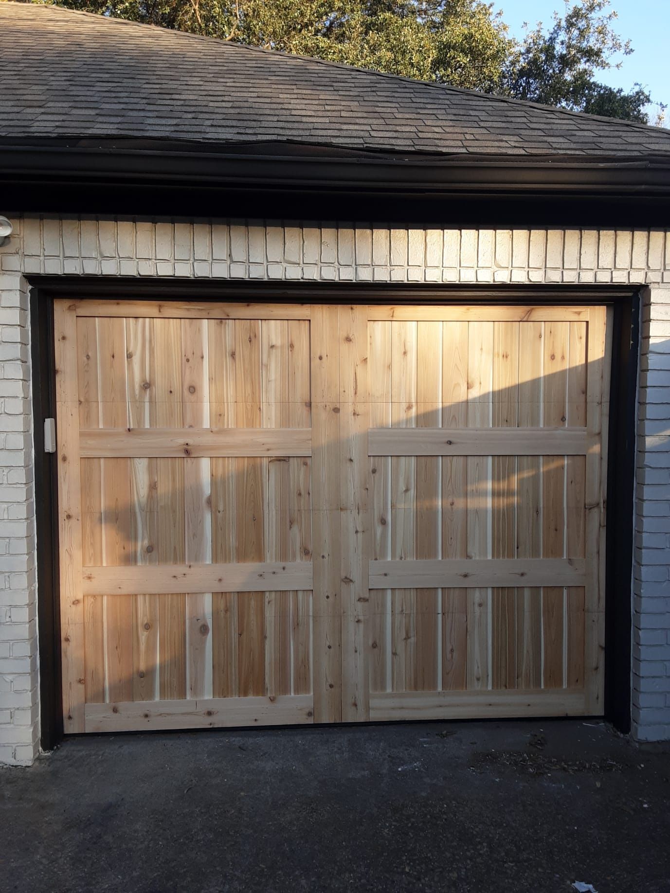 A double garage door made of light-colored vertical cedar planks with horizontal trim, set in a white brick wall.