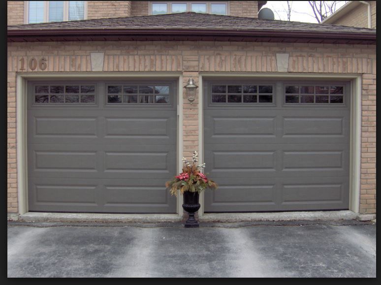 Two gray, paneled garage doors on a brick house, with a small potted plant sitting on the ground between them.