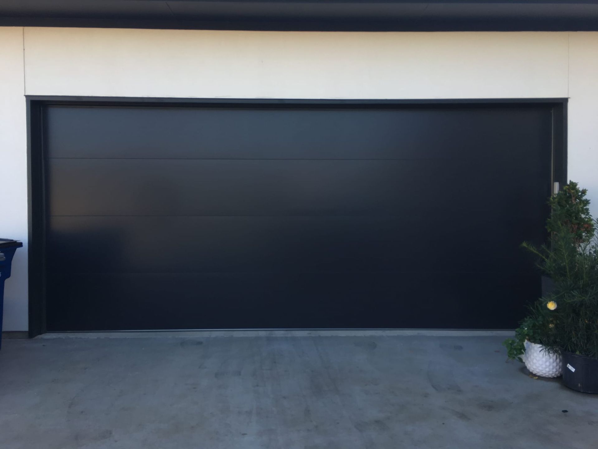 A wide, flat-panel garage door in matte black, installed on a white building exterior next to a potted plant.