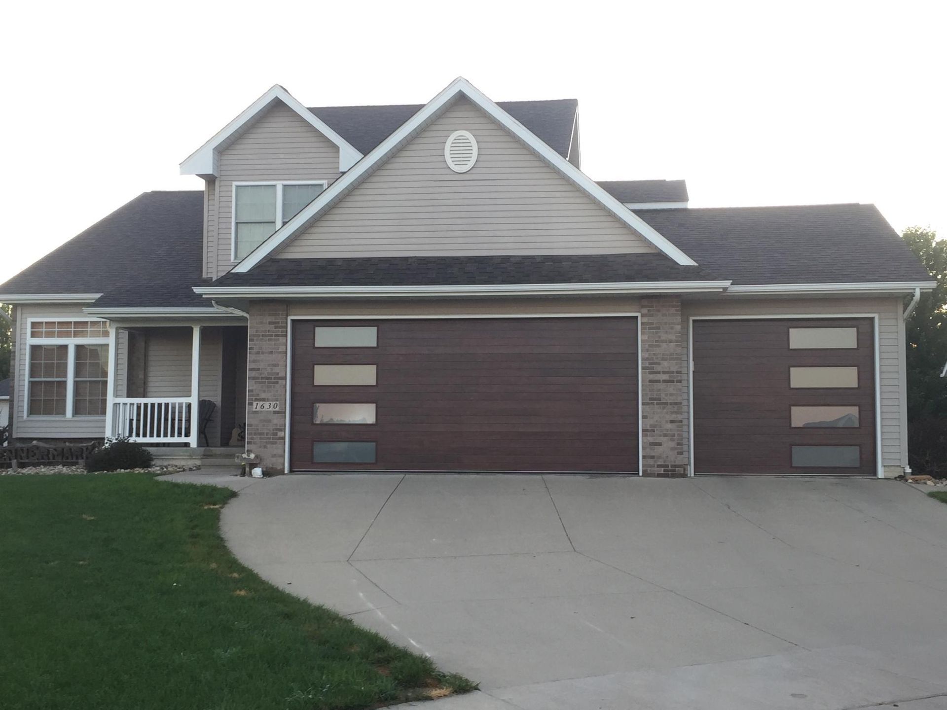 A two-story suburban house with beige siding, dark brown garage doors with horizontal glass panels, and a front porch.