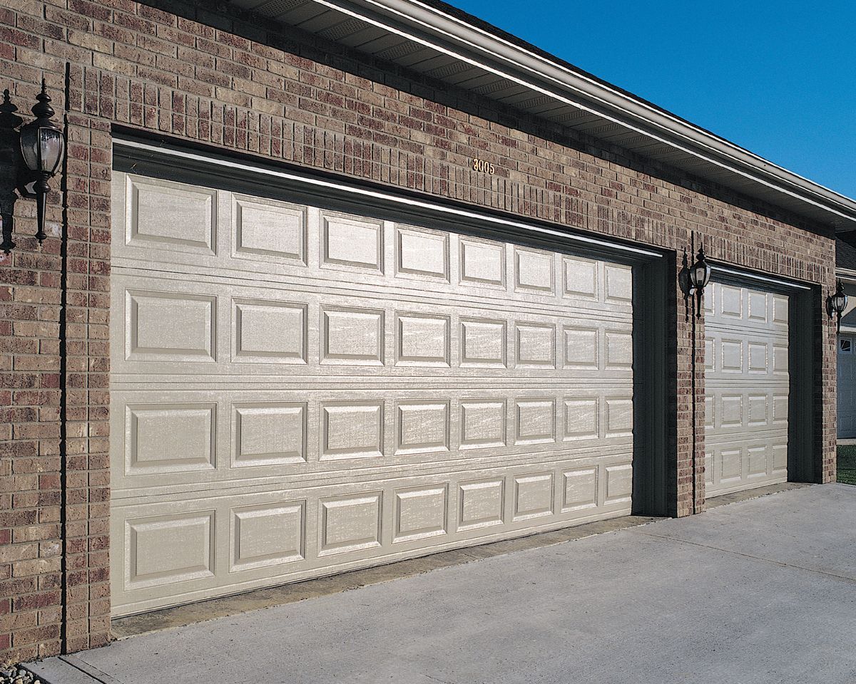 A beige two-car garage with raised panels, framed by brown brick, under a clear blue sky.