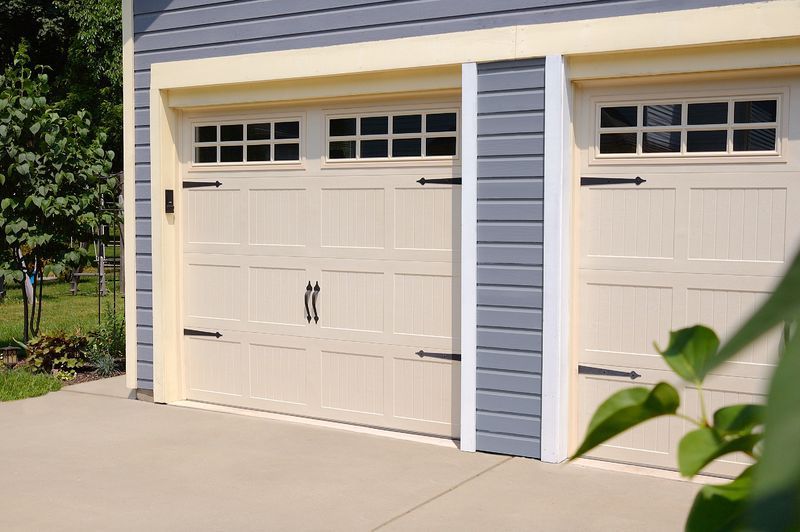 A two-car garage with cream-colored, paneled doors featuring decorative black hinges, handles, and windows.