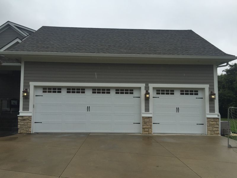 A gray garage with a dark shingled roof, stone accents at the base, and two white garage doors with top-row windows.