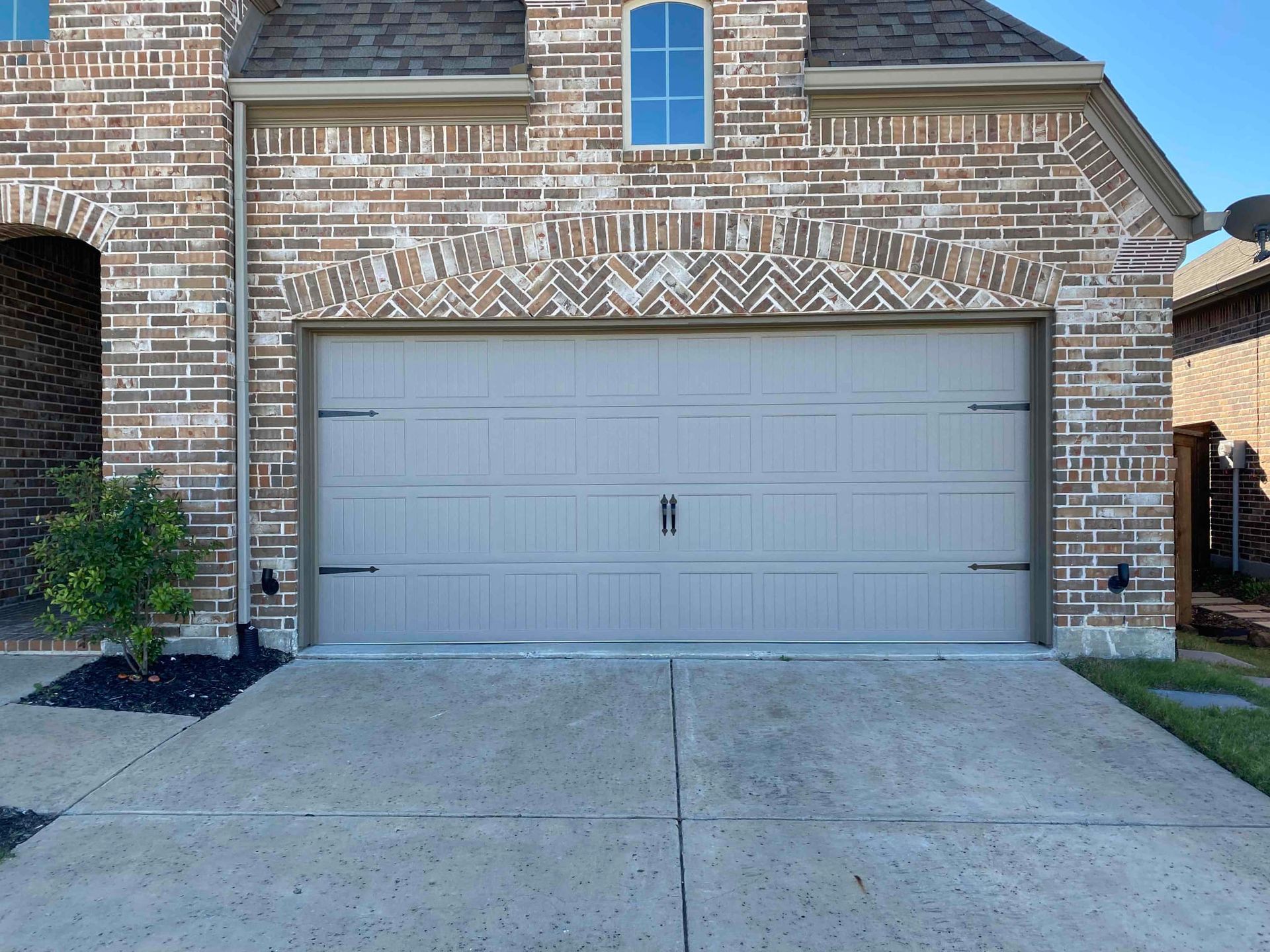 A tan, modern garage door set in a decorative brick wall with a herringbone arch above it and a concrete driveway.