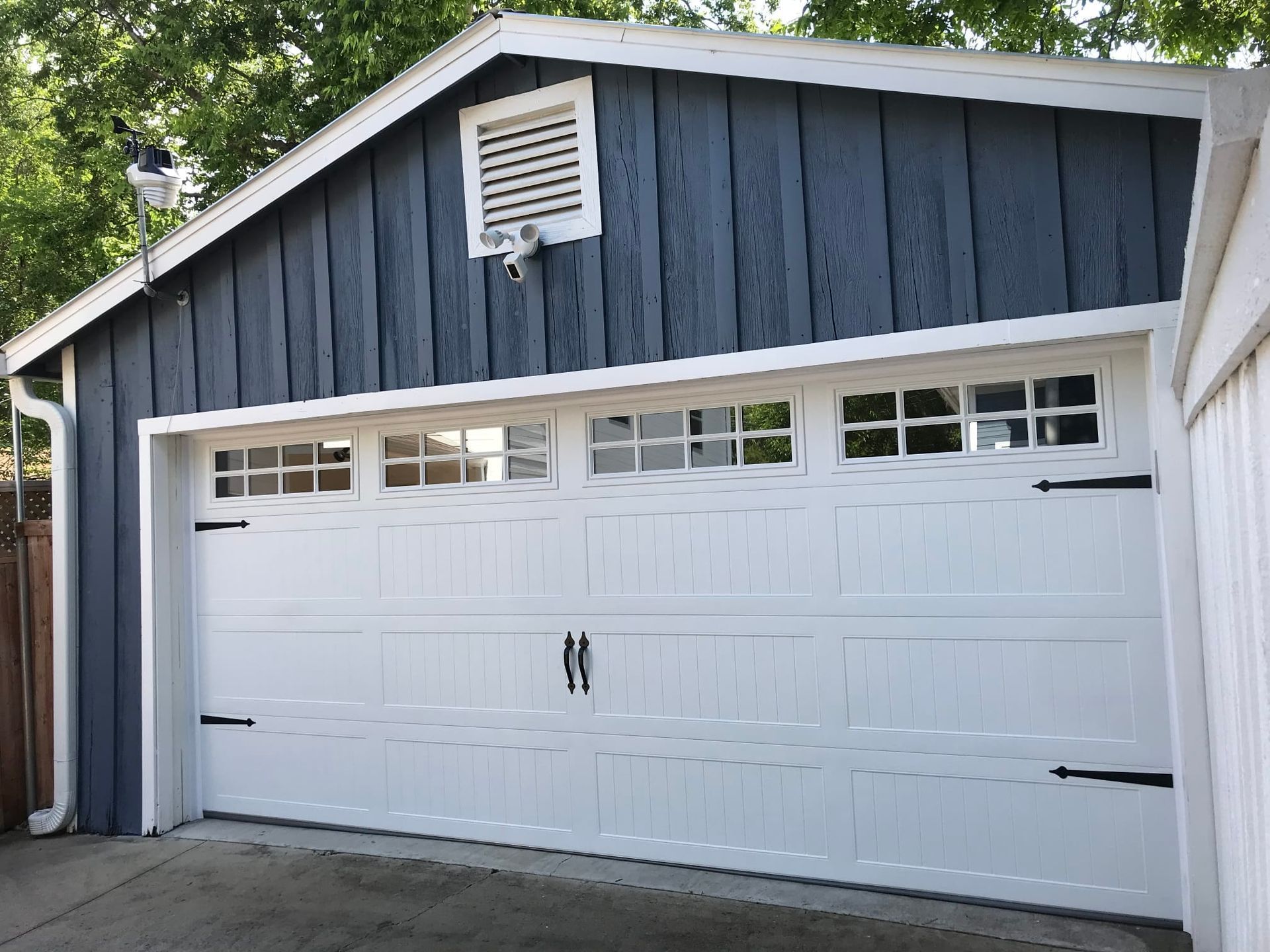 A white, four-panel residential garage door with window inserts, set beneath a dark blue, vertical-sided garage exterior.