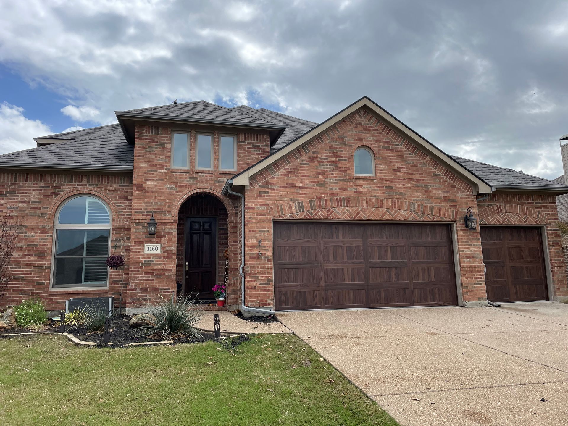 A single-story brick house with a multi-car garage, dark brown doors, and an arched entry under a cloudy sky.