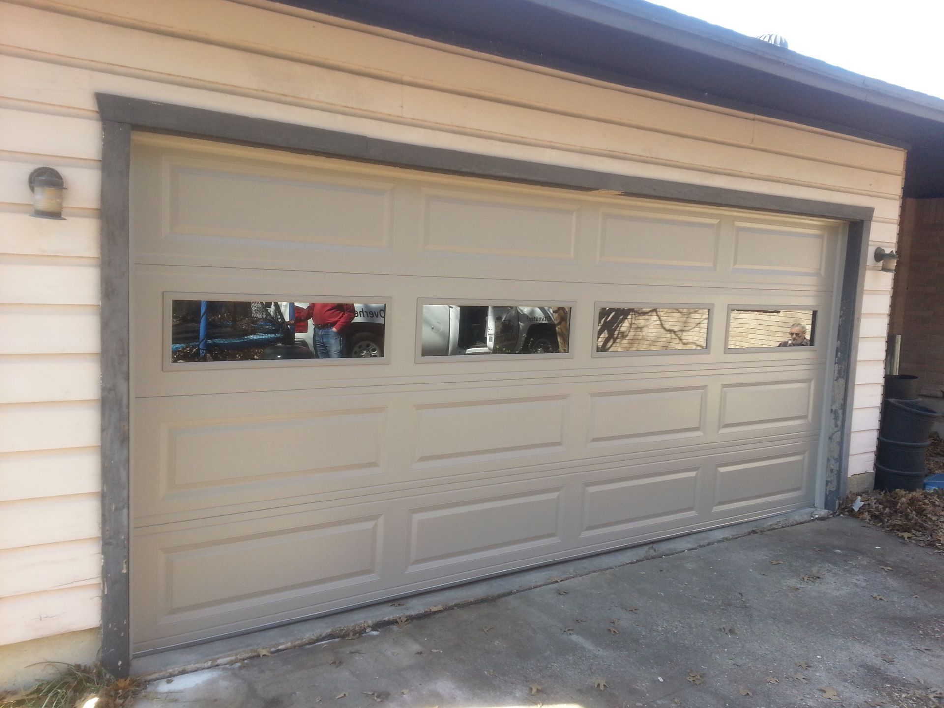 A taupe, four-panel residential garage door with a row of four horizontal windows installed on a house with light siding.