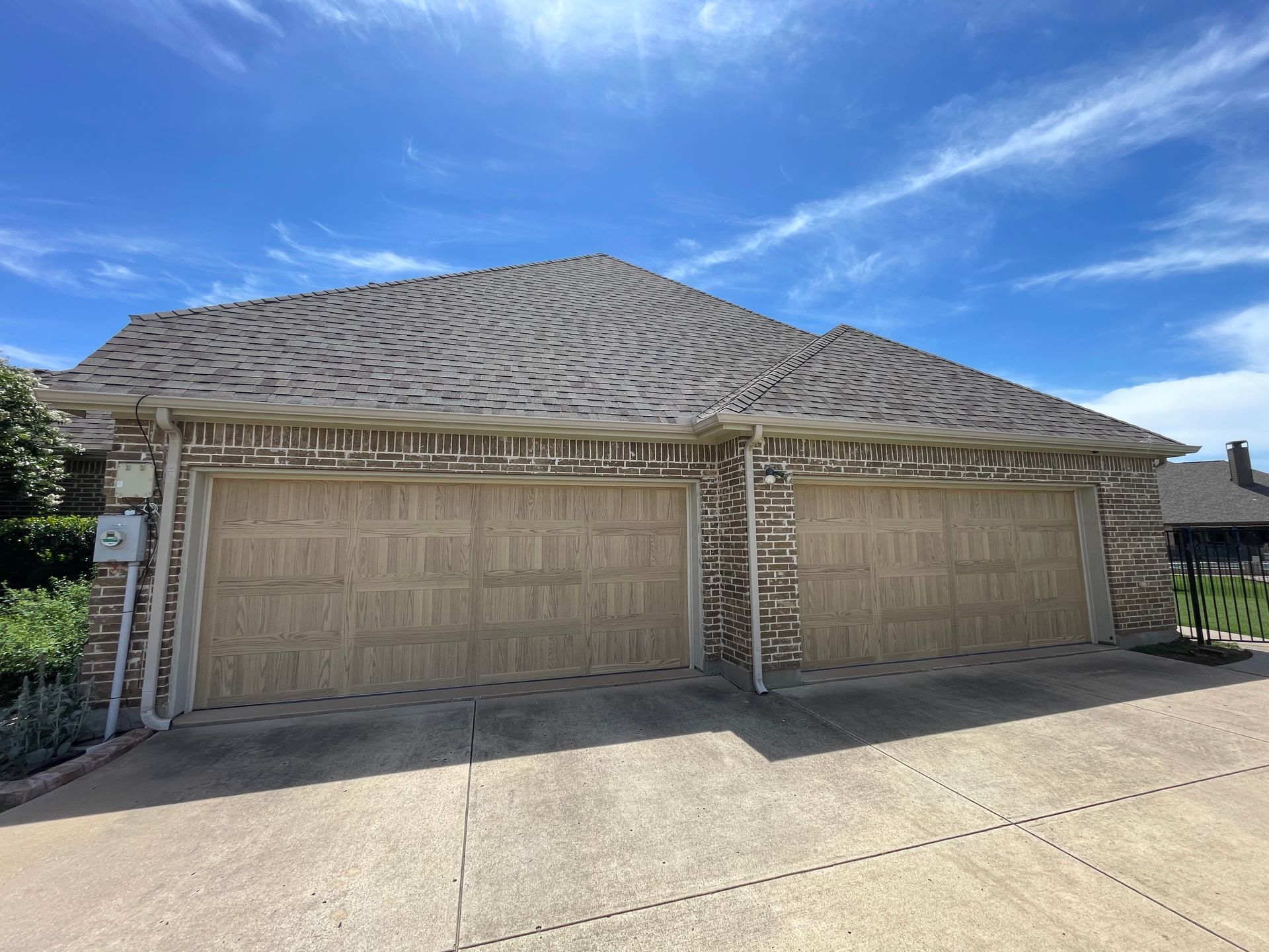 A tan brick house with a two-car garage under a dark shingled roof against a bright blue sky.
