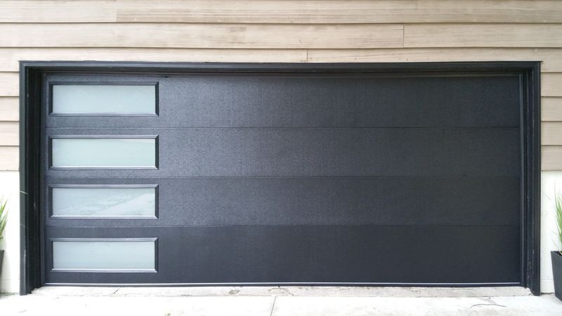 A modern black garage door with a vertical column of four frosted glass rectangular windows on the left side.