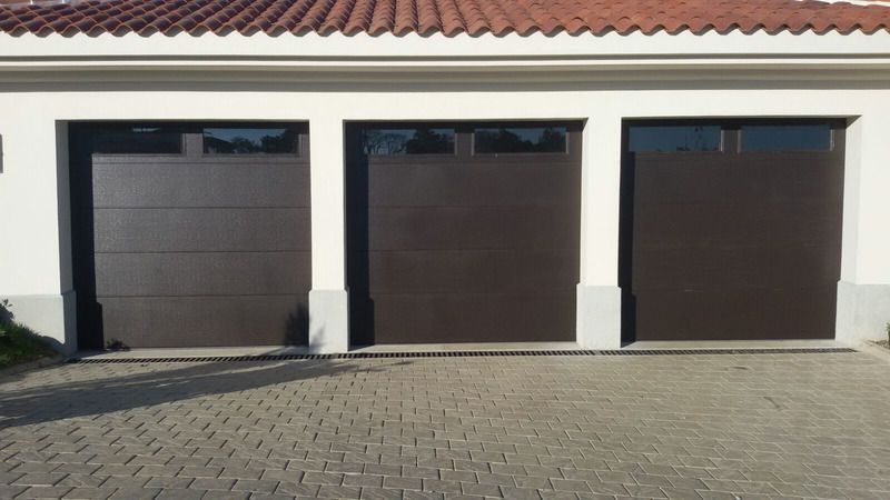 Three dark brown garage doors with rectangular windows along the top, set into a light-colored building with a tiled roof.