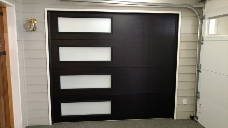 A modern black garage door with a vertical row of four frosted glass windows on the left side, set against gray siding.