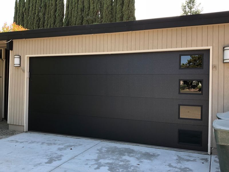 Modern black garage door with four vertical stacked windows on the right side, installed on a beige house.