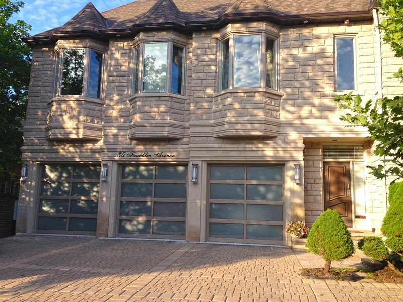 A large, multi-story stone house with three modern frosted-glass garage doors and a paved driveway at sunset.