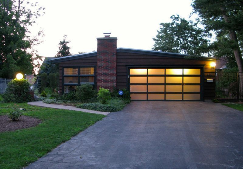 A dark-toned, mid-century modern ranch house with a brick chimney and a large, glowing, multi-paneled glass garage door.