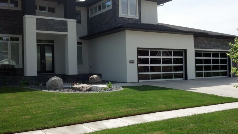 A modern house with a green lawn, stone accents, white walls, and two contemporary glass-paneled garage doors.