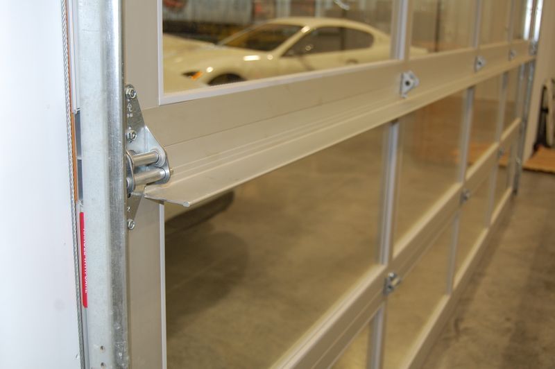 Close-up of a hinge and roller mechanism on the side of a modern aluminum-framed glass garage door inside a garage.
