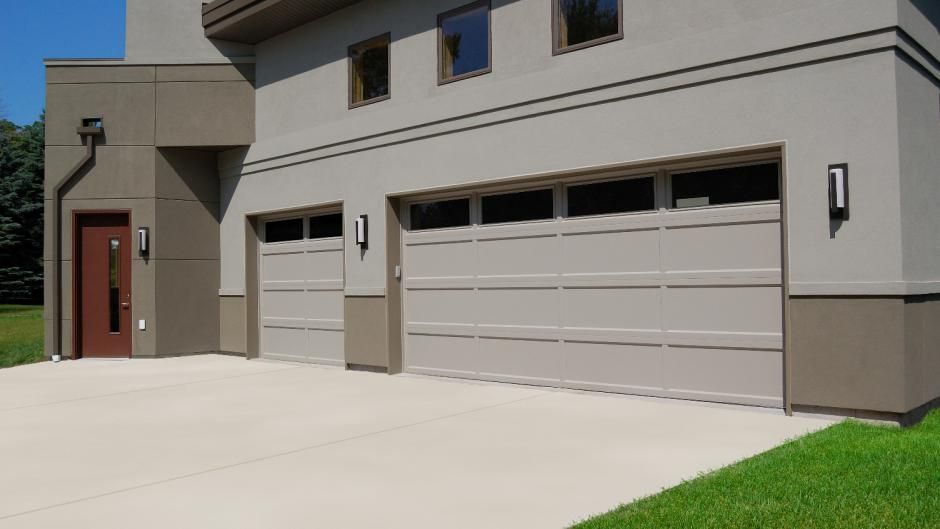 A modern two-story building with a brown door, a single-car garage, and a double-car garage under a clear blue sky.
