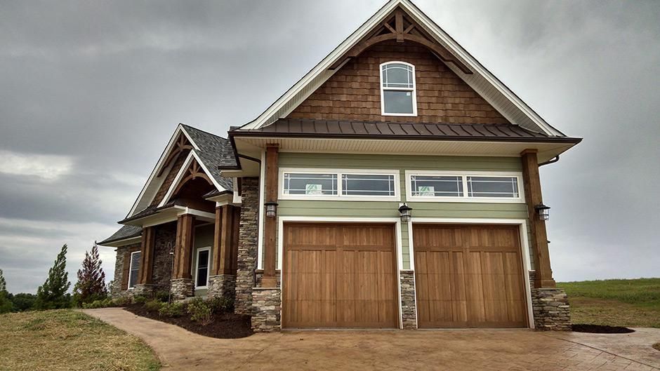A two-story, Craftsman-style home with wood garage doors, stone accents, and a cedar-shingled gable under a cloudy sky.