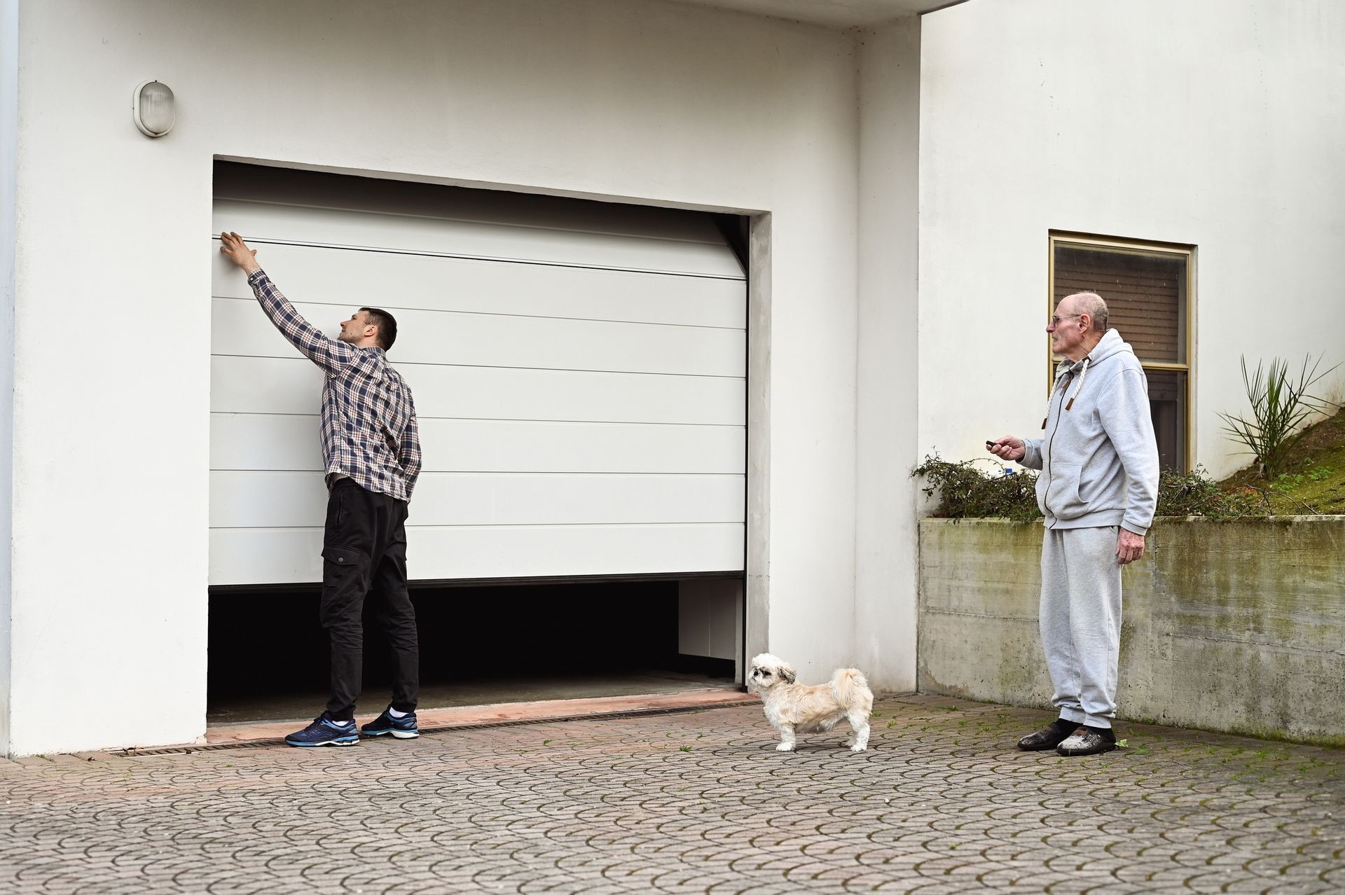 Man opening garage door, older man nearby holding a remote, small dog in front.