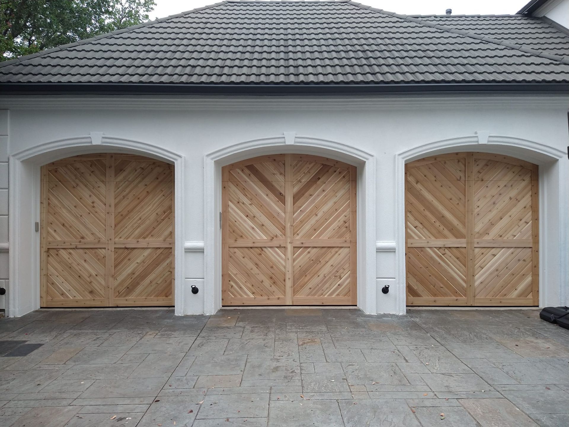 Three arched wooden garage doors with diagonal paneling set into a white stucco building with a shingled roof.
