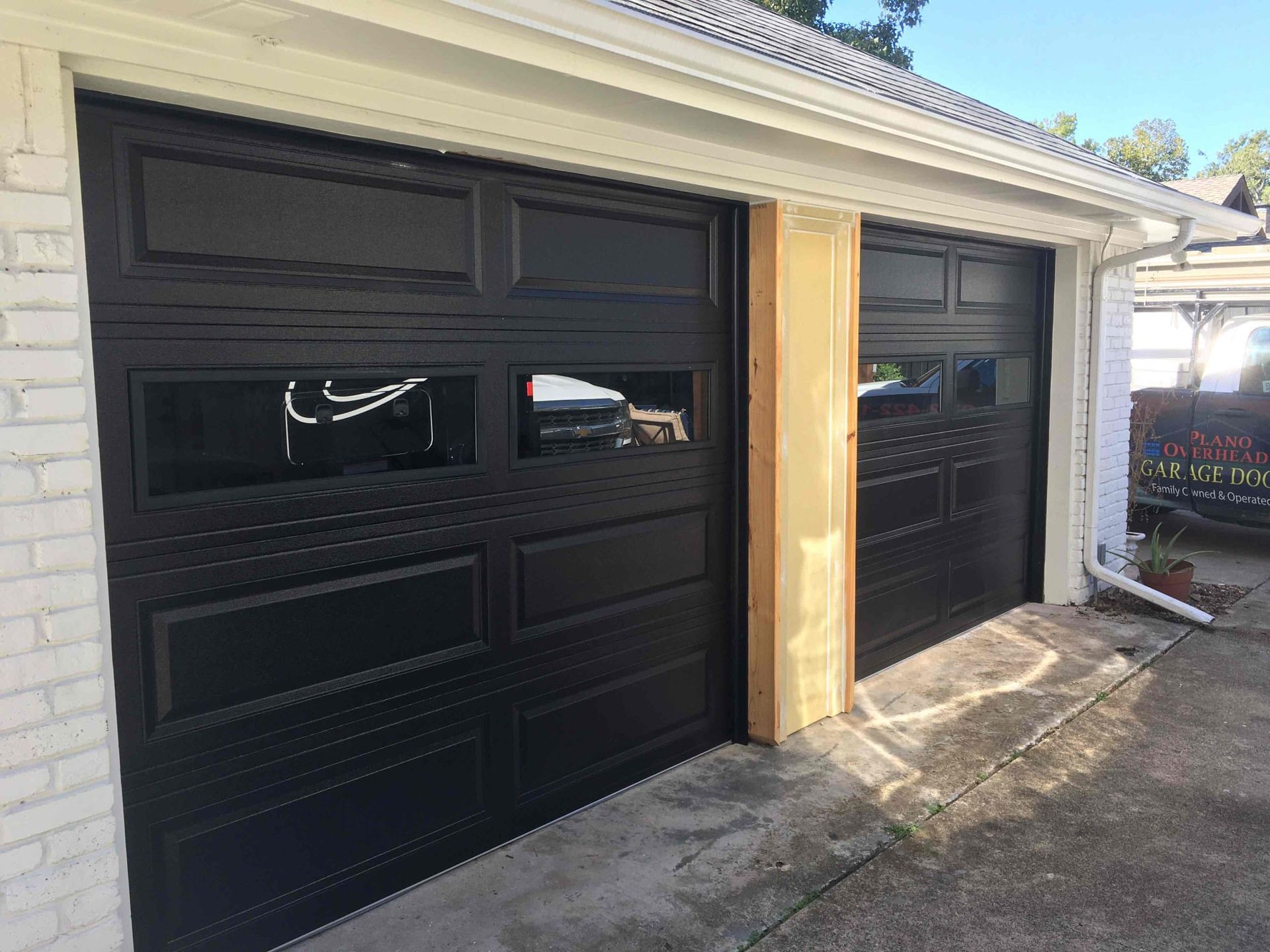 Two black, paneled residential garage doors with horizontal windows installed side-by-side in a white brick building.