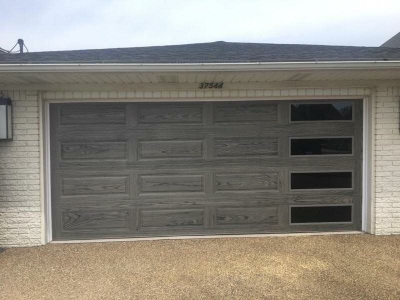 A gray wood-grain garage door with four rectangular windows vertically aligned on the right side, set in a brick wall.
