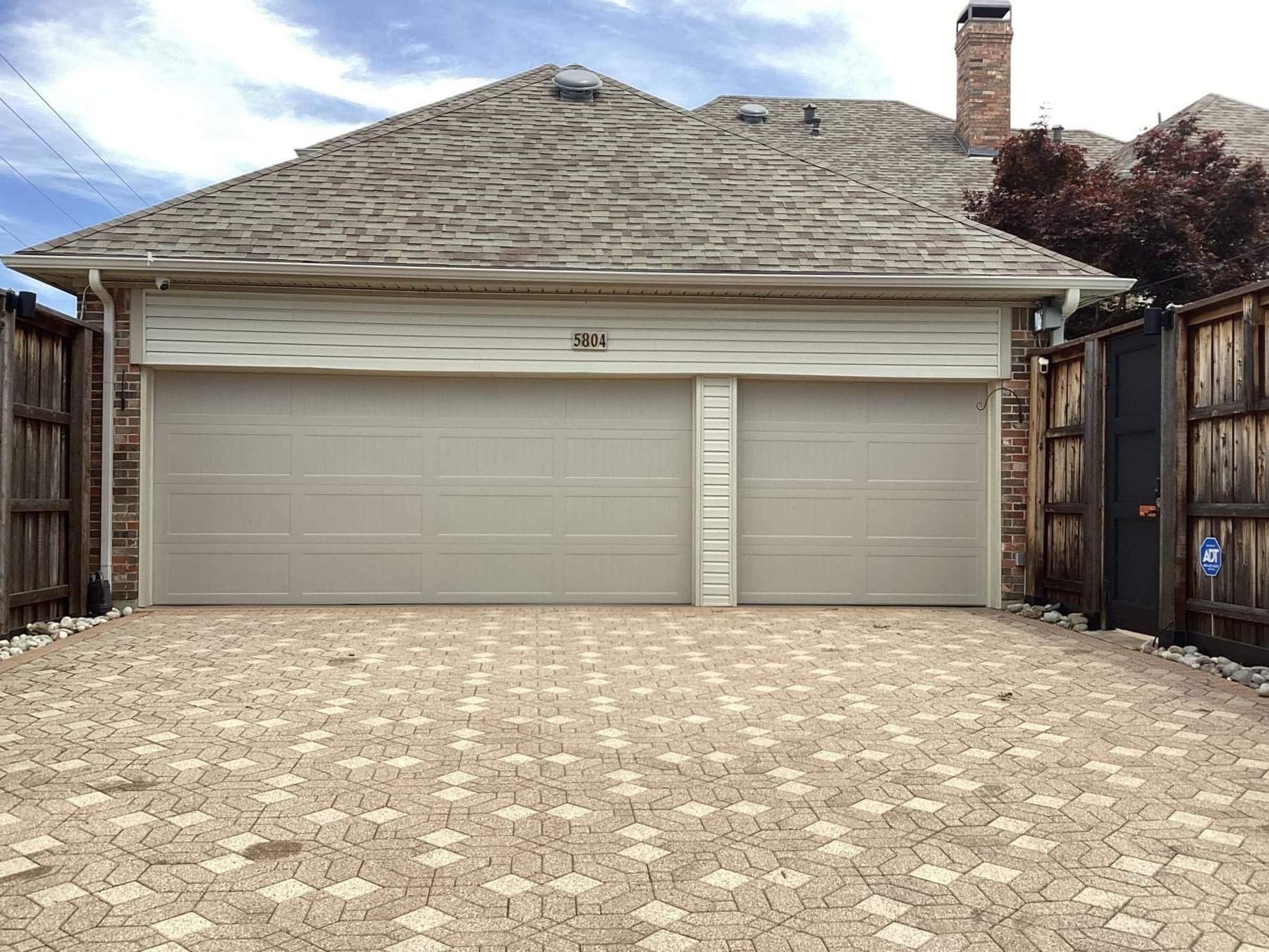 A residential garage with two light-colored doors and a decorative paved driveway in a suburban setting.