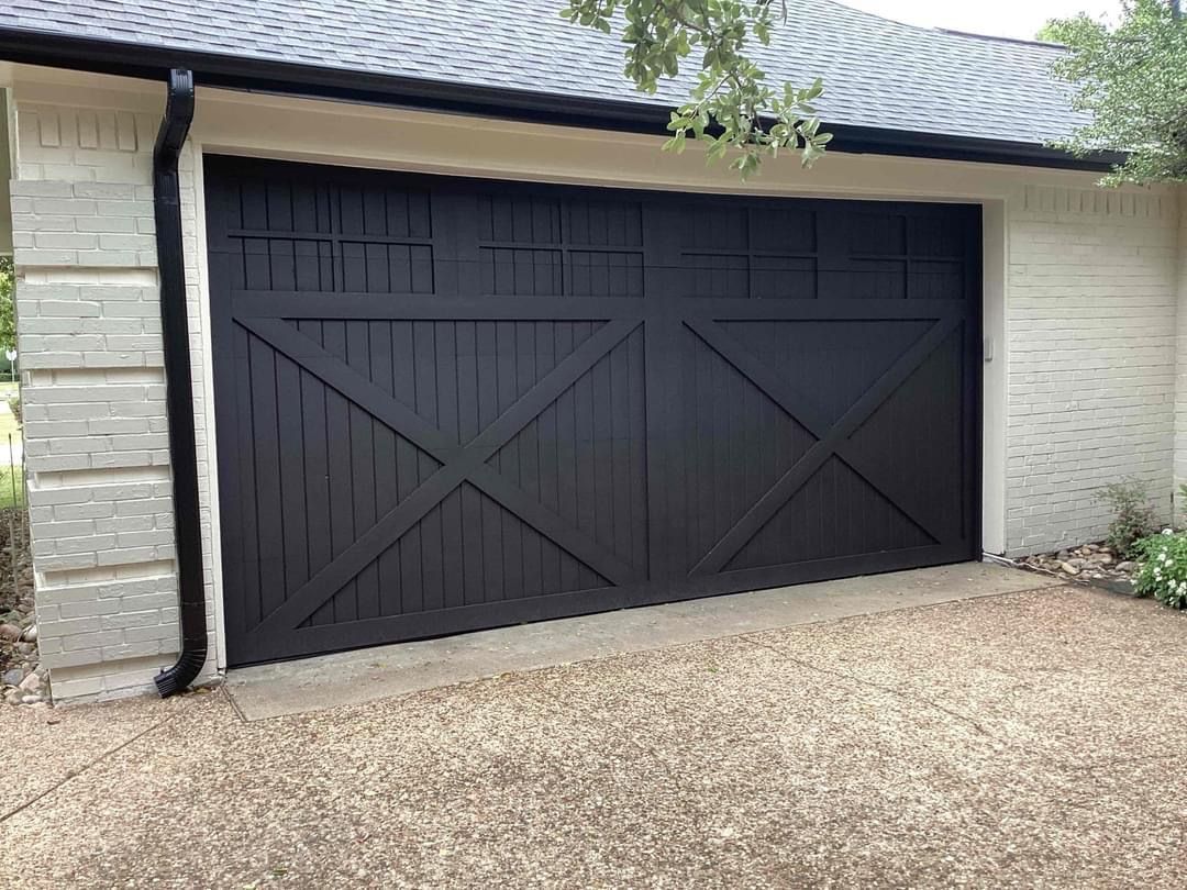 Black carriage-style garage door with X-shaped cross-bracing and horizontal window panels, set in a light stone building.