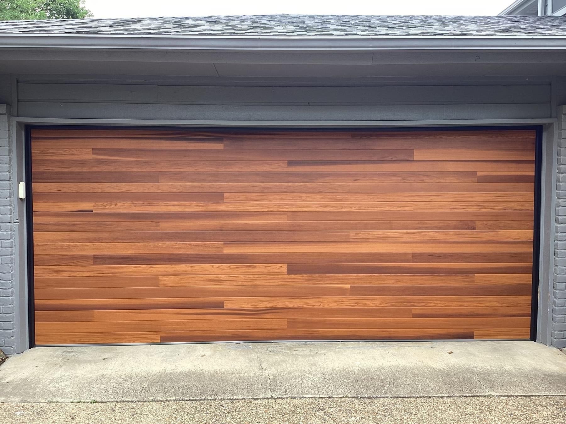 A modern garage door with horizontal planks of warm, multi-toned wood grain, framed by gray siding and brick.