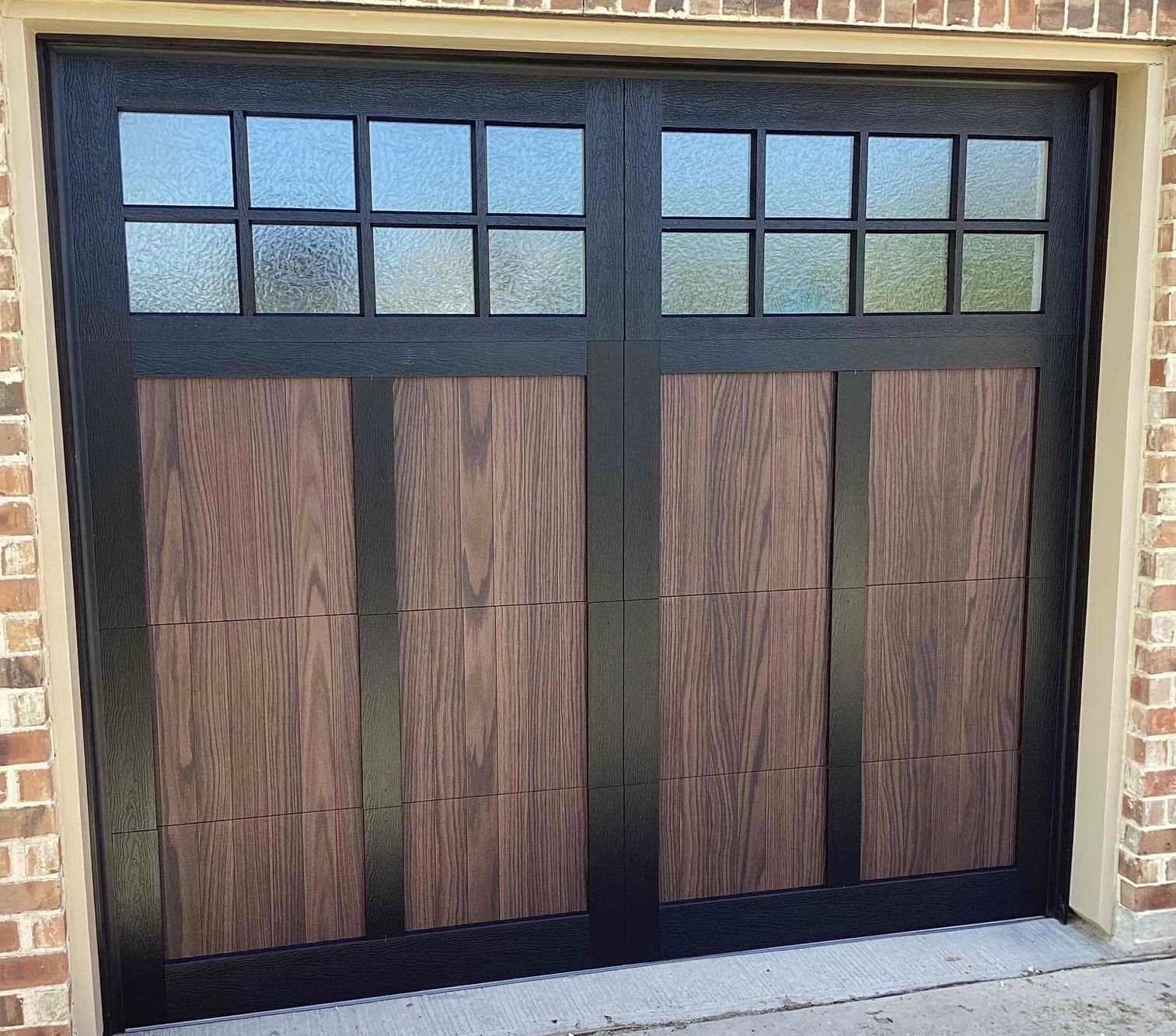 A garage door with black frames, wood-grain panels, and two rows of glass window panes, set in a brick wall.