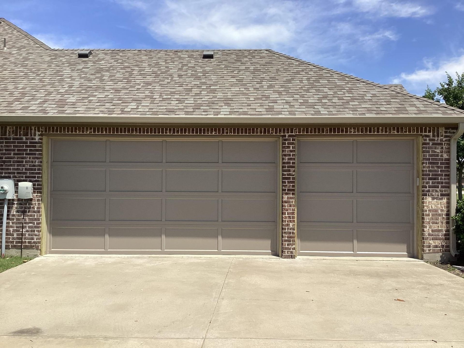 A beige two-car garage with horizontal panels under a brown roof and brick exterior walls.