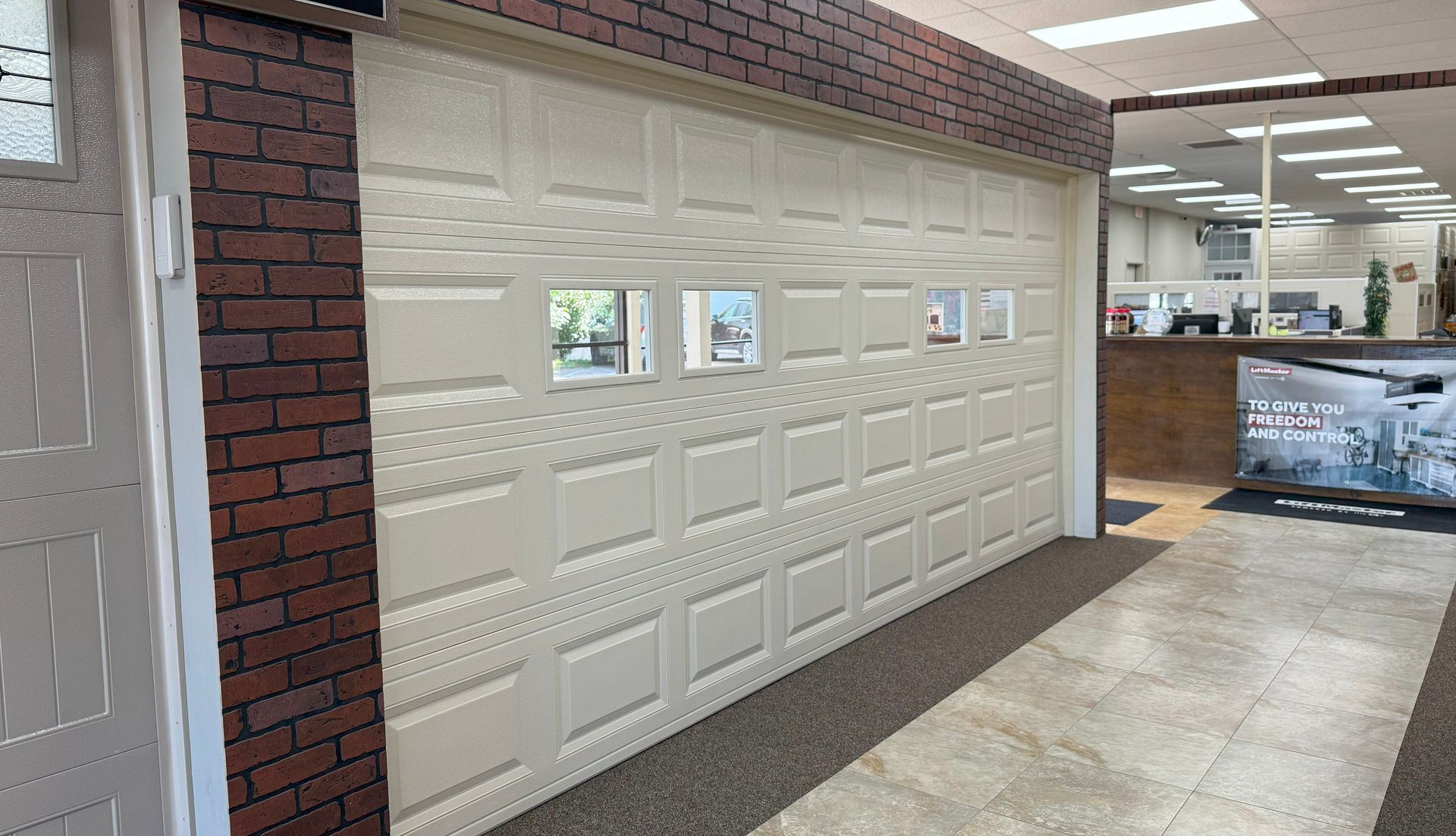 Beige garage door with windows, on display in a showroom with brick wall and office in background.