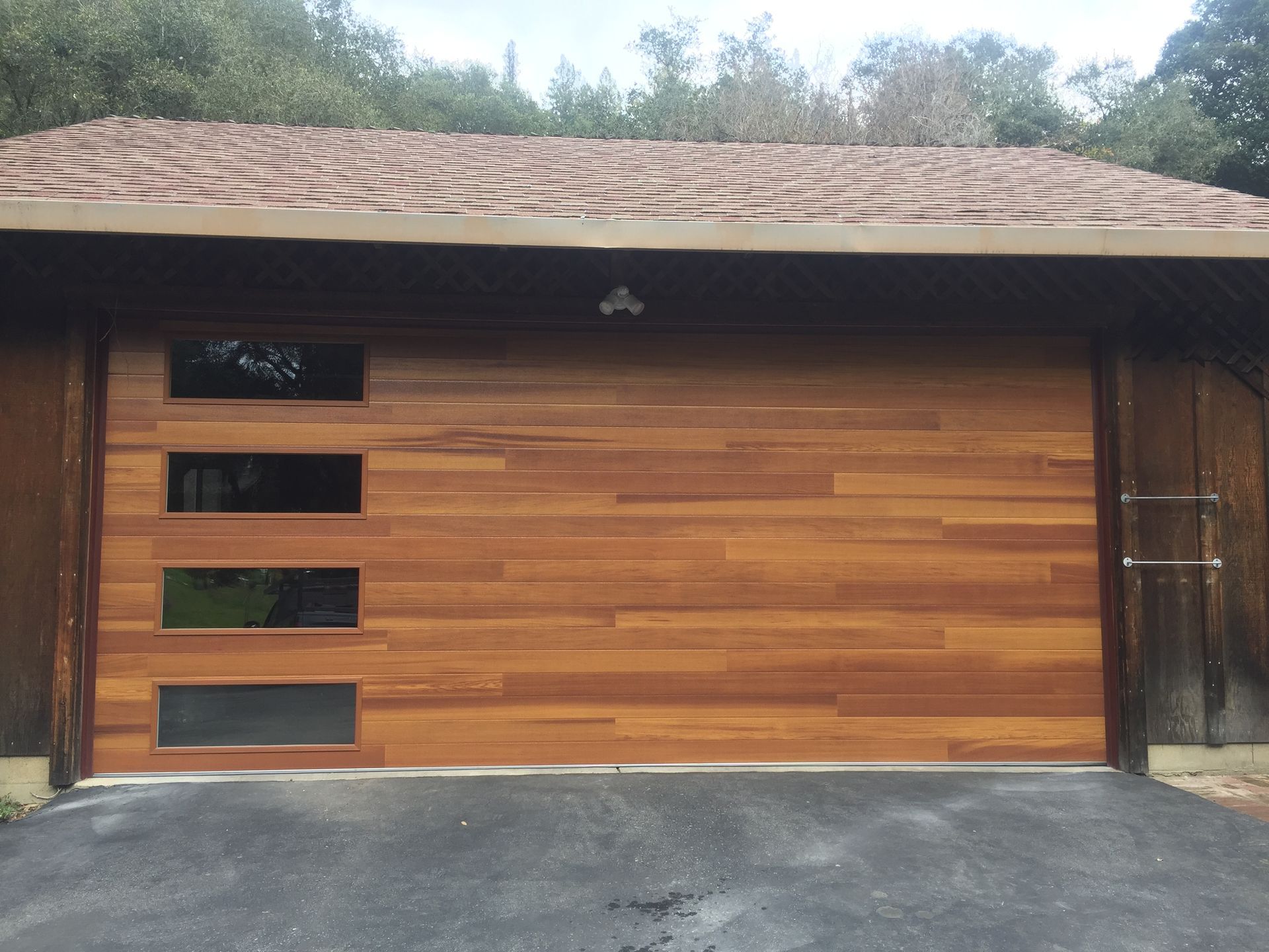 Modern wood-paneled garage door with a vertical row of four rectangular glass windows on the left, set in a paved driveway.
