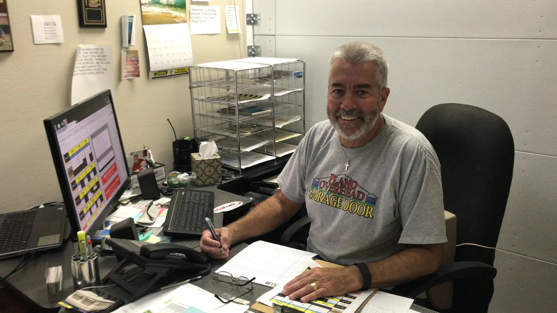 Man at desk, smiling, in front of a computer.  He holds a pen, gray hair, and wears a grey t-shirt.
