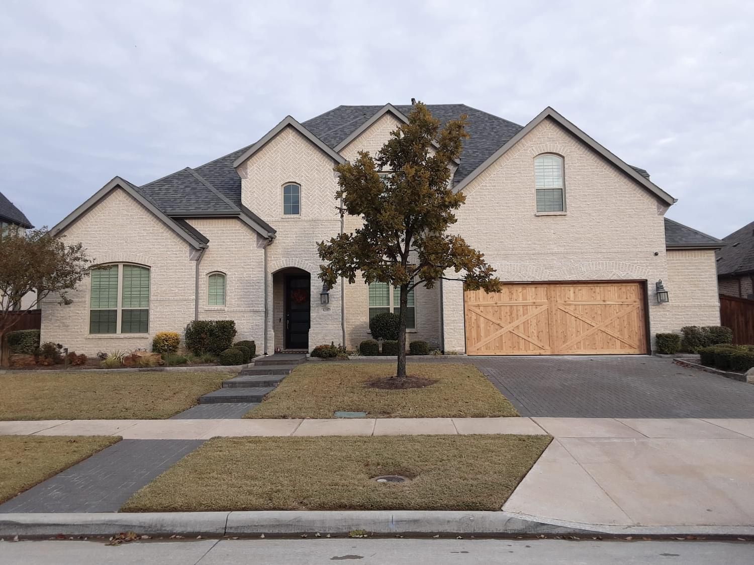 Two-story beige brick house with a gray roof and a wooden garage door on a cloudy day.