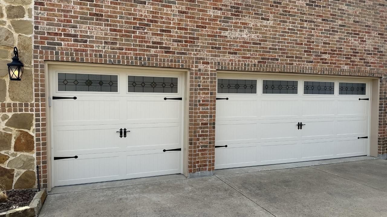 Two white garage doors with decorative black hinges, handles, and windows on a brick exterior house.