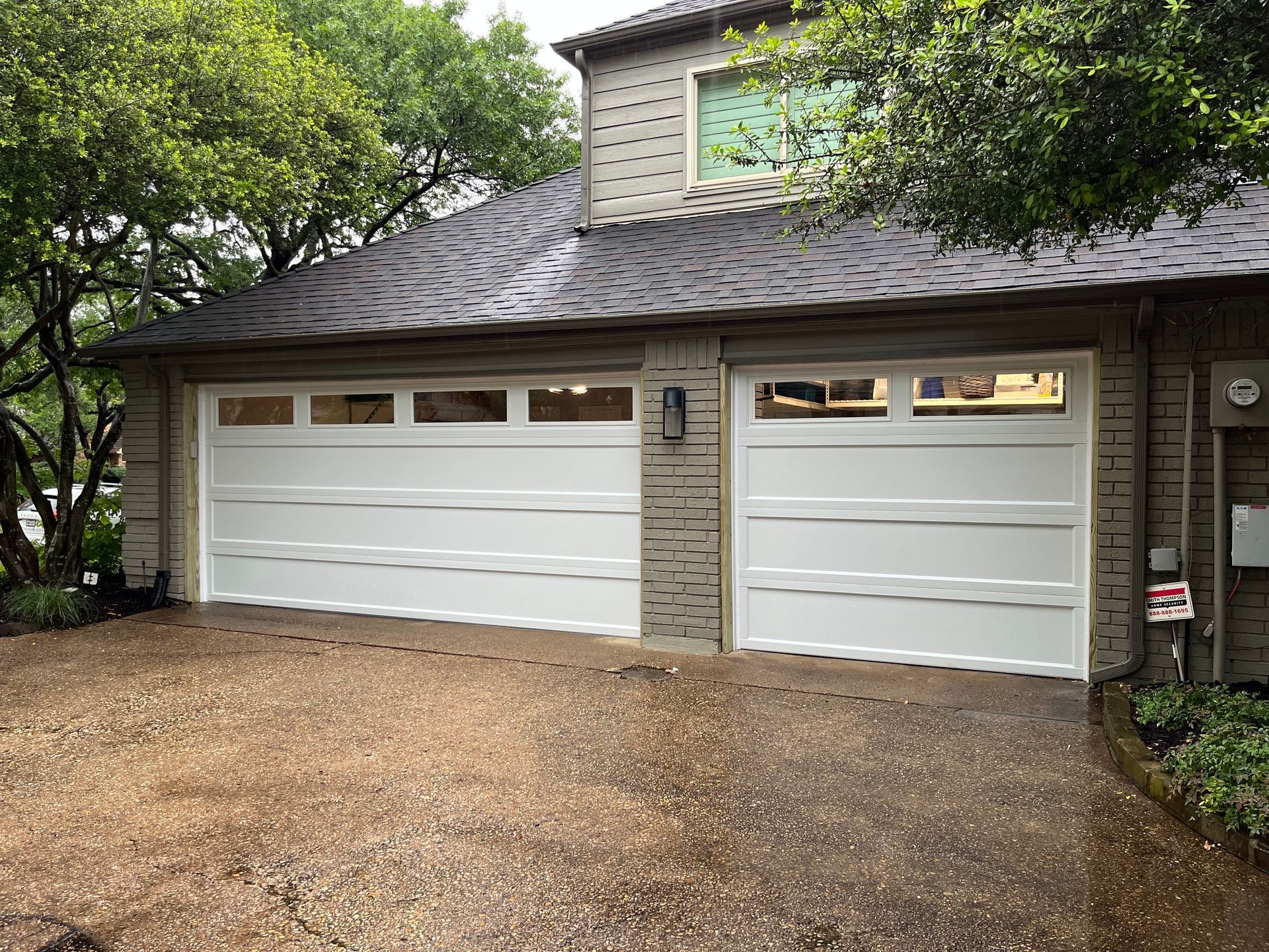 White garage doors with top-row windows on a brick house front with a gravel driveway and surrounding trees.