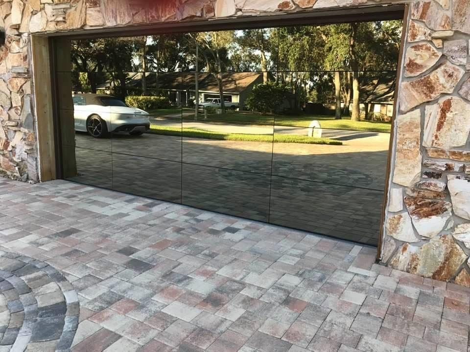 A modern mirrored garage door reflecting a house and driveway, set within a rustic stone frame on a paved area.