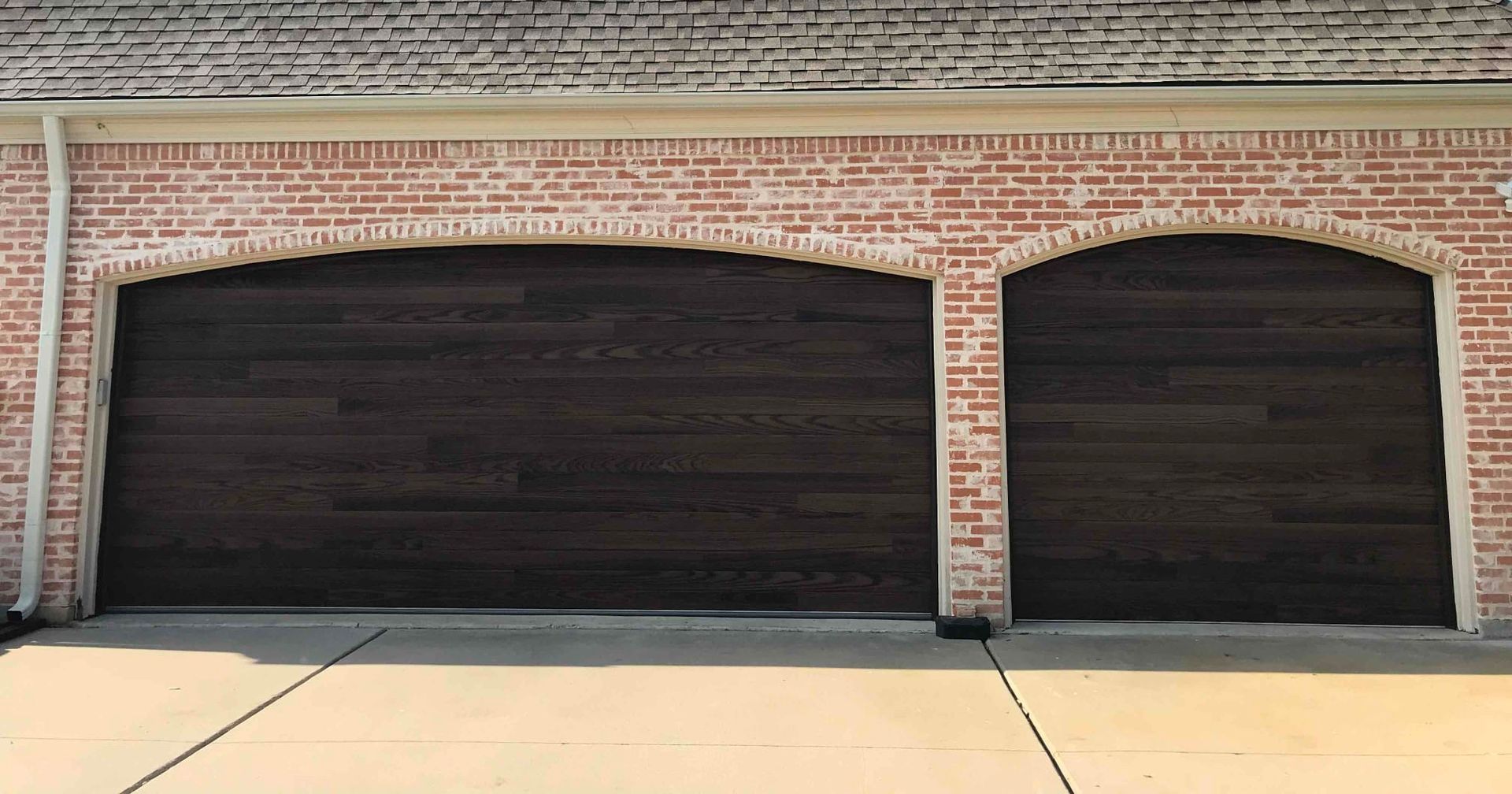 Two dark-stained wood garage doors with arched tops set within a red brick wall above a concrete driveway.