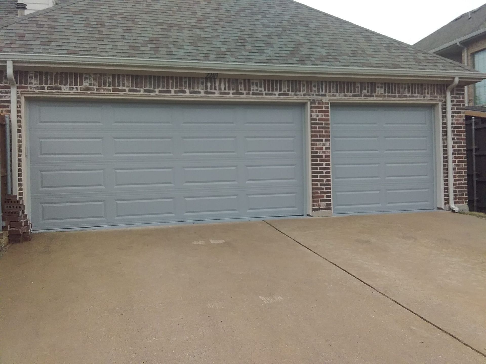 A grey two-car garage with a large and a small door set in a red brick house with a shingled roof.
