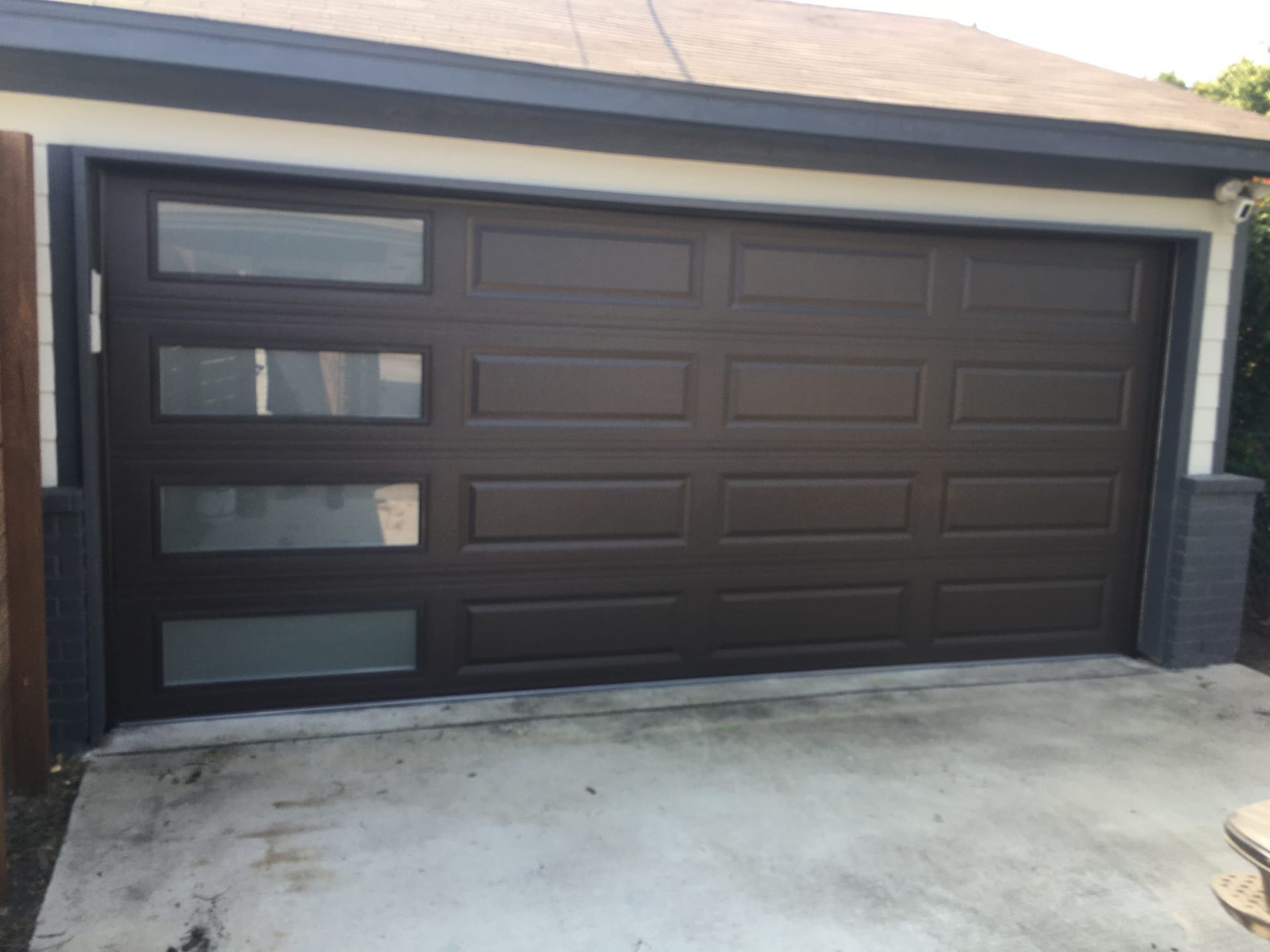 Dark brown garage door with a vertical row of four frosted glass windows on the left side, set into a light-colored home.