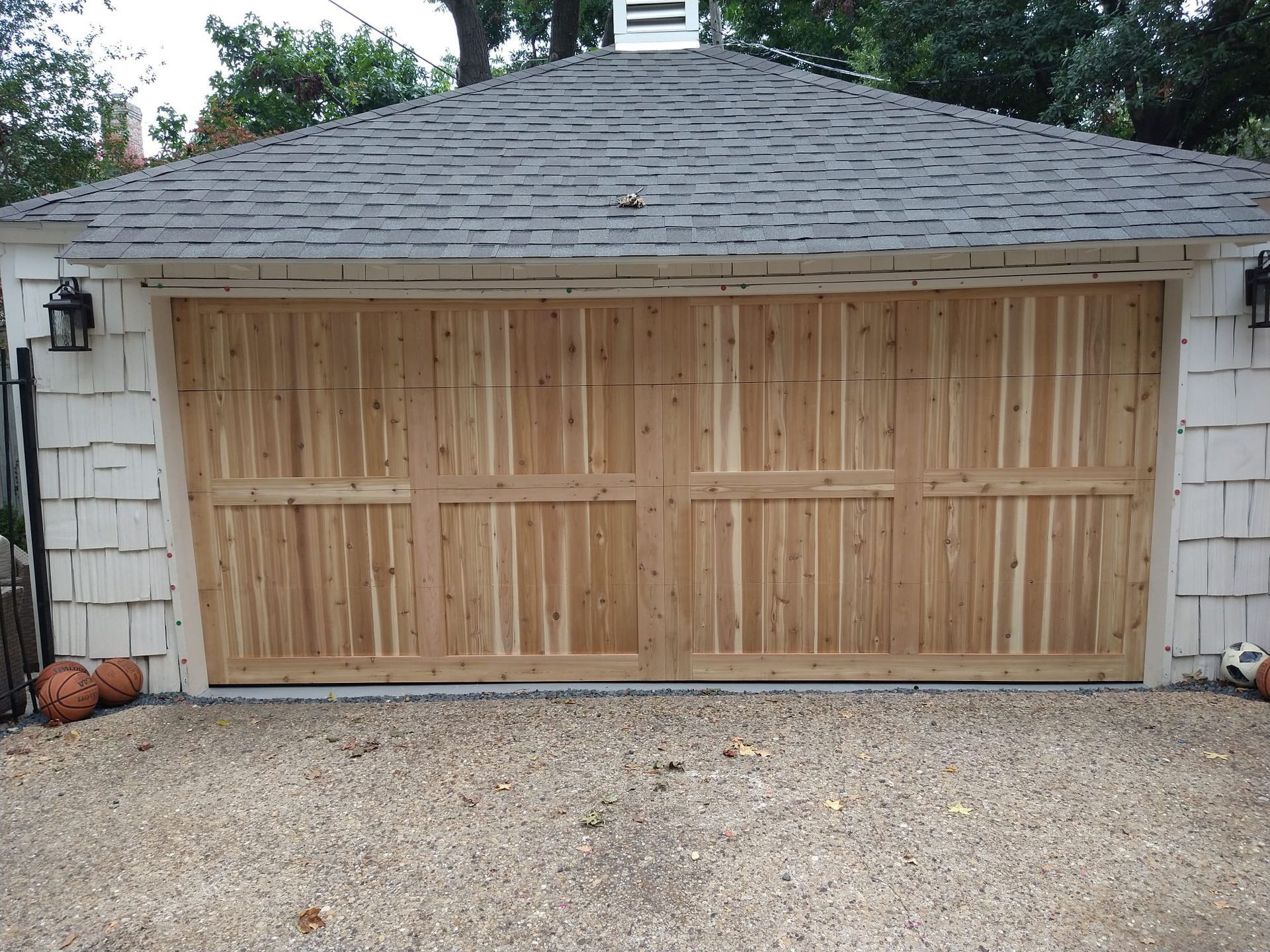A wooden carriage-style garage door on a white shingled building with a shingled roof, set above a gravel driveway.