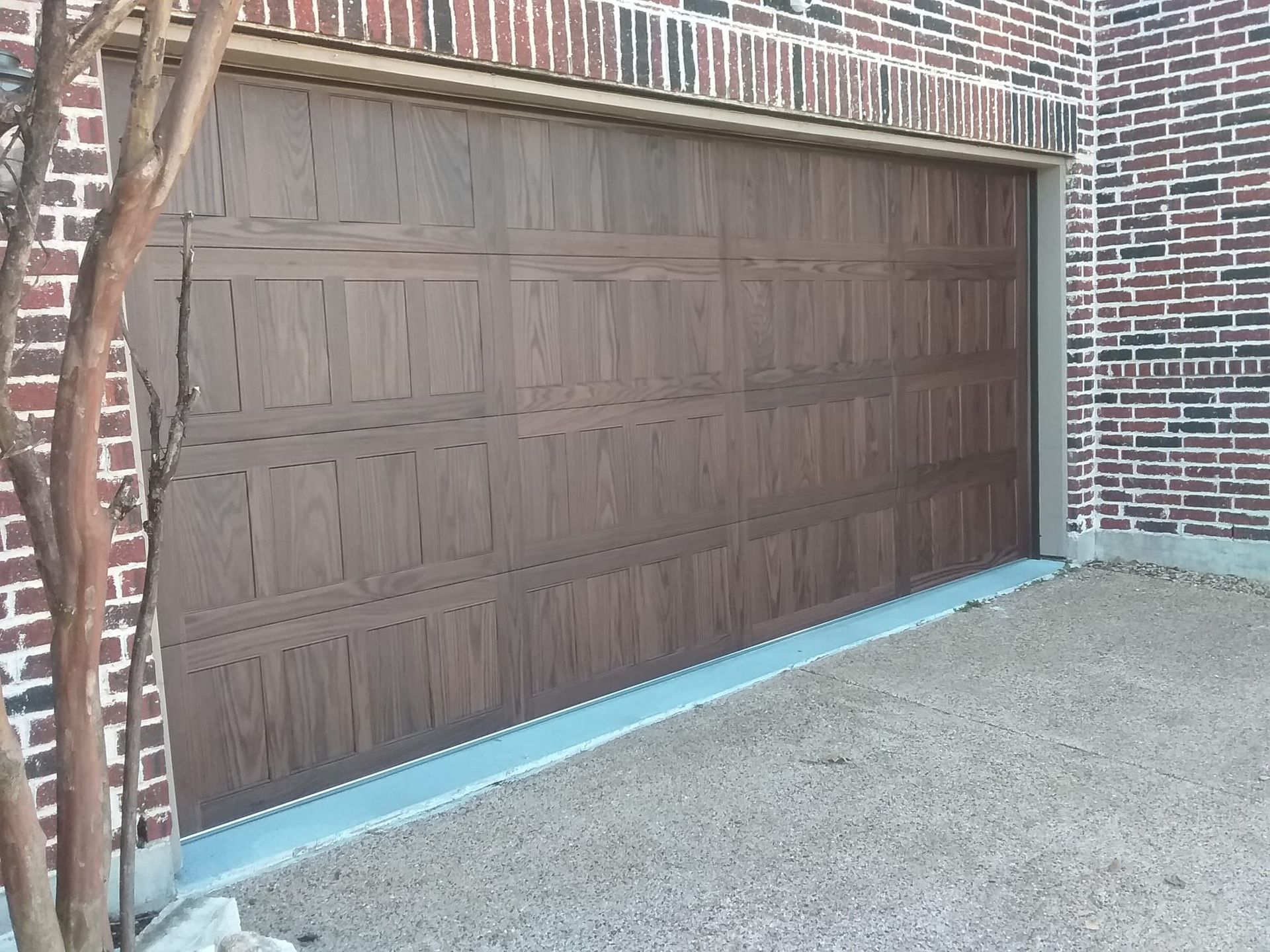 A dark brown garage door with a paneled wood-grain design, installed in a brick house exterior.