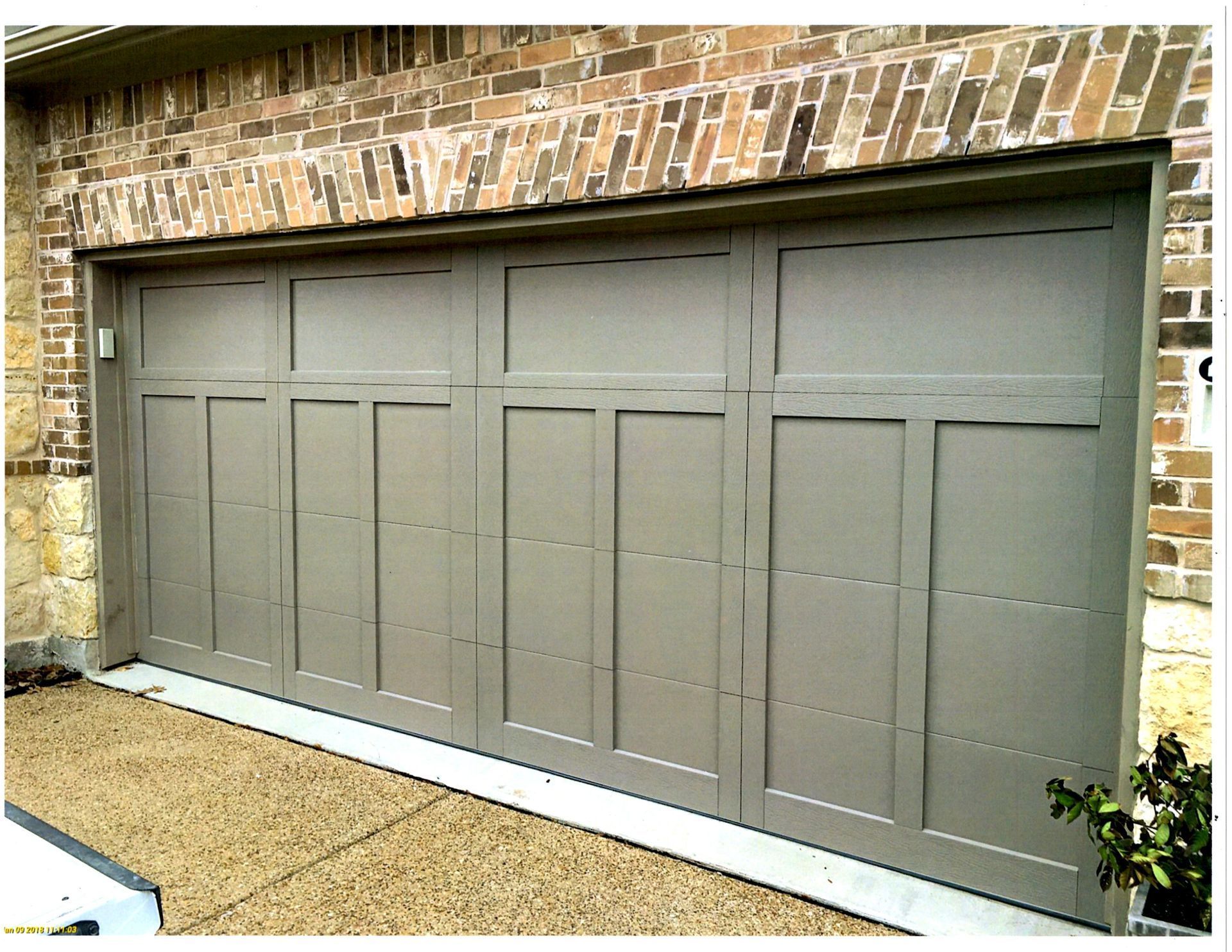 A closed, gray, multi-panel garage door installed in a brick home exterior with a light-colored concrete driveway.