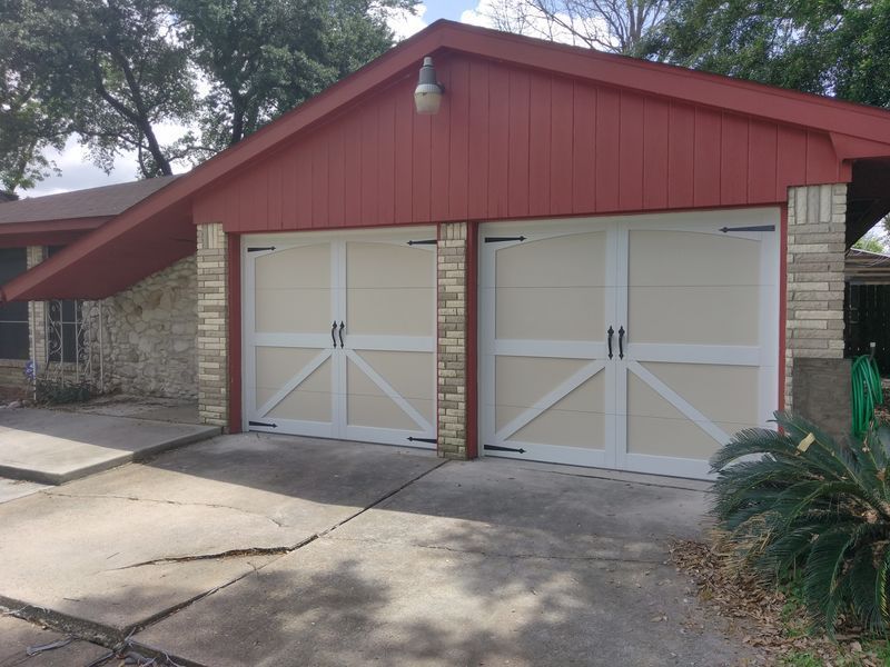 A tan two-car garage with white cross-buck door trim and black hardware, set in a stone and red-sided home exterior.